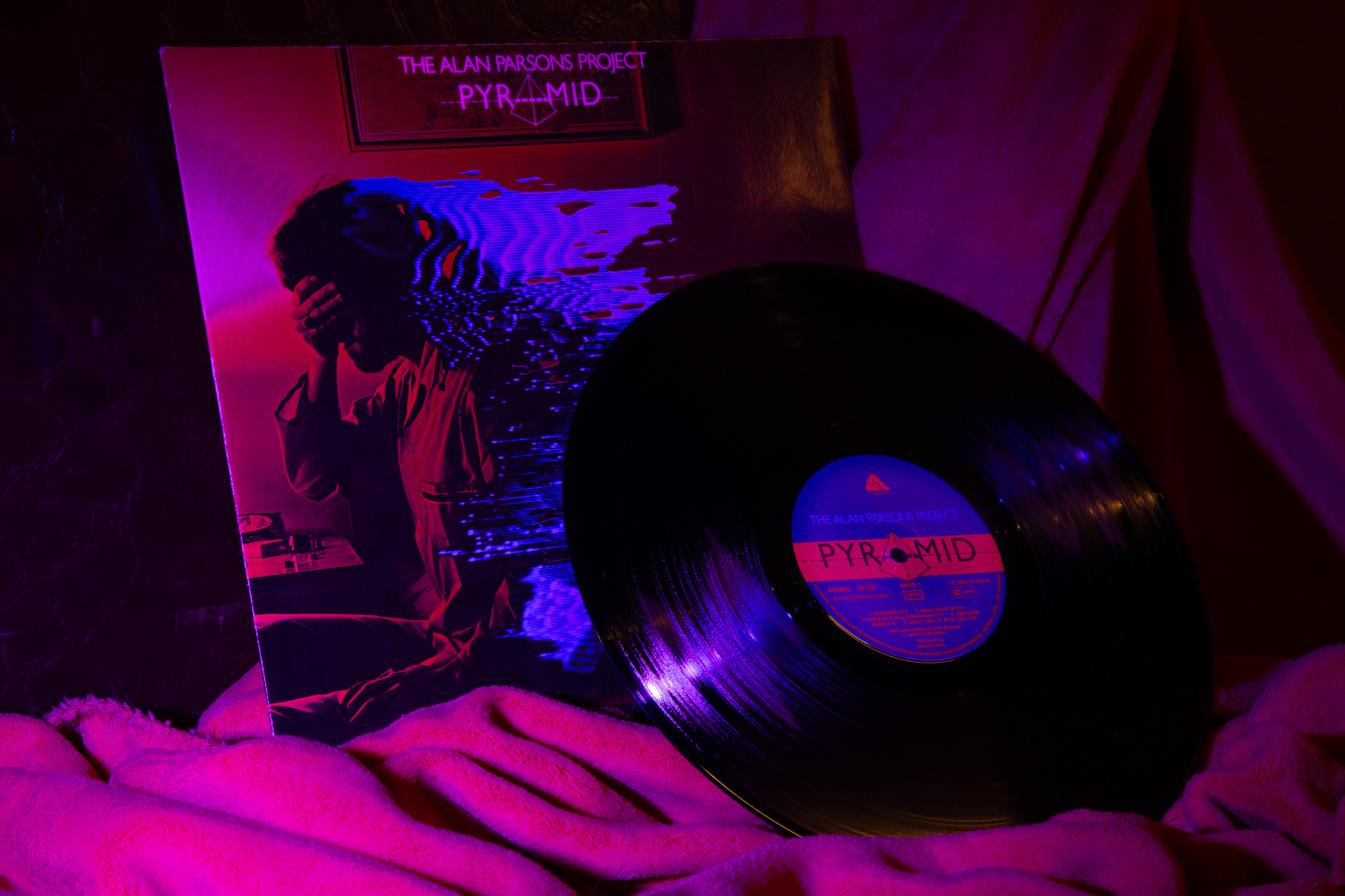 The vinyl of The Alan Parsons Project Pyramid is put on some soft fabric resembling sand its sleeve right behind it. It's lit in a magenta hue makeing the blue of the sleeve and the vinyl label pop.
